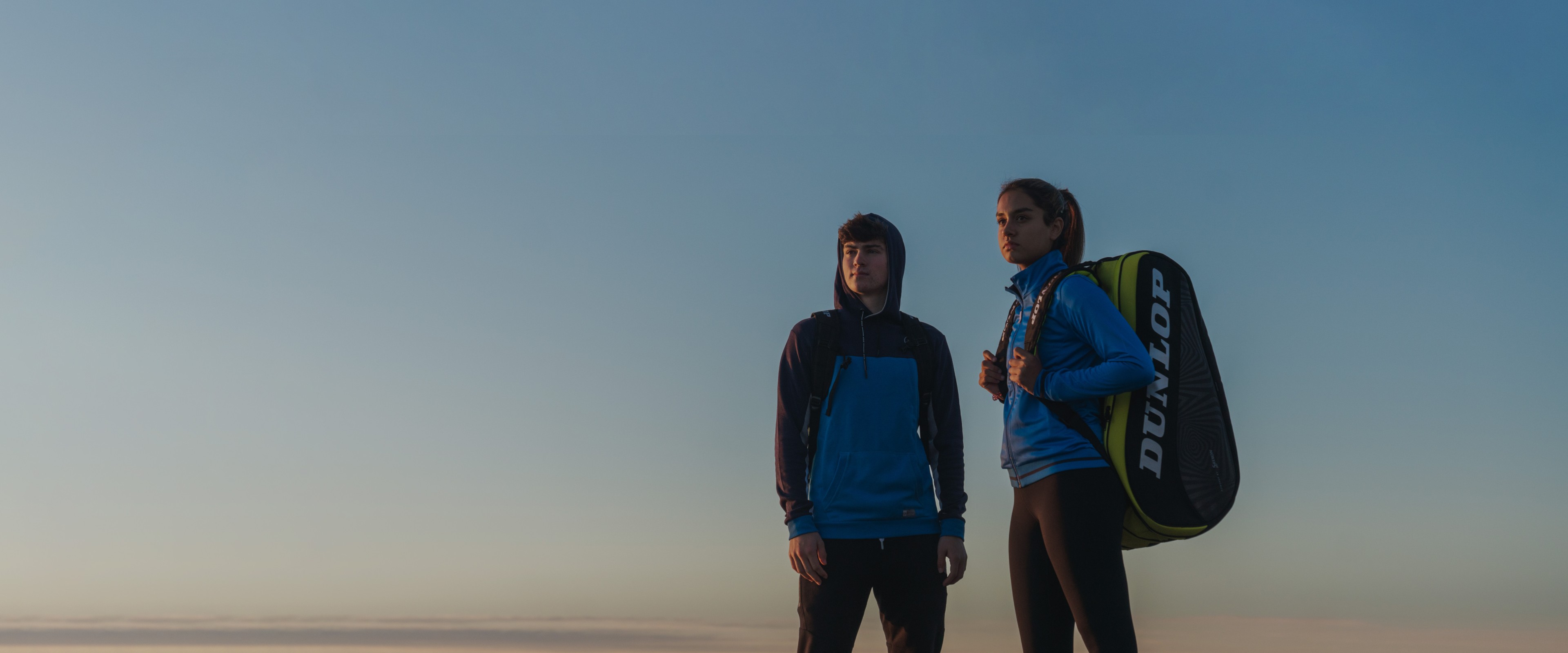 A man and woman standing on a beach wearing a Dunlop Sports backpack